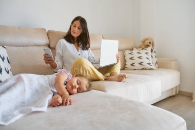 Dizüstü bilgisayarı ve akıllı telefonu karıştıran olumlu genç bir kadın. Oturma odasında bebek kaçırma ve serbest proje üzerinde çalışırken kanepenin yanında oturuyor.