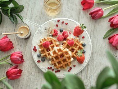 Tabakta çeşitli meyvelerle süslenmiş gözlemeler kırmızı lalelere karşı bal kavanozu ve masada bir bardak kahve.