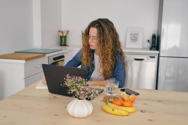 Yoğunlaşmış kadın girişimci, günlük giysiler içinde ahşap masada meyvelerle oturuyor ve Netbook 'a göz atarken not defterine not alıyor ve evdeki mutfakta uzak bir proje üzerinde çalışıyor.