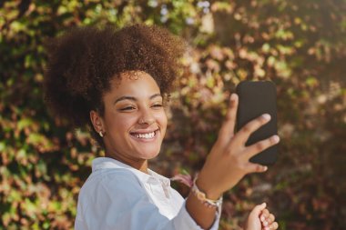 Kıvırcık saçlı Afro-Amerikalı bir kadın. Parkta güpegündüz dişlek bir gülümsemeyle akıllı telefondan kendi portresini çekiyor.