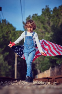 Kot pantolonlu çocuk, raylar arasında ezilmiş taşların üzerinde yürürken ve dalgalanan Amerikan bayrağını tutarken aşağıya bakıyordu.