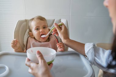 Pembe önlüklü sevimli bir kız bebek yüksek sandalyede oturuyor ve evde tanınmayan bir kadının yardımıyla kaşıktan yemek yiyor.