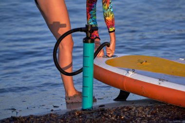 Anonim kadın sörfçü mayo giyip gün batımında dalgalanan mavi suyun yanında SUP tahtalarını pompalıyor.