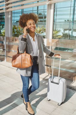 Tüm vücudu kıvırcık saçlı, şık giyinen, bavulla şehirde dikilen ve cep telefonuyla konuşan bir kadın.