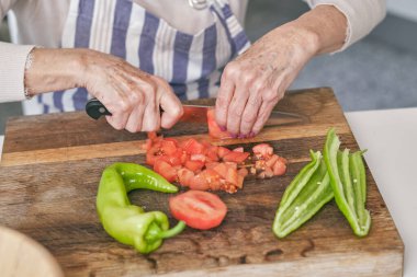 Anonim bir kadın, açık mutfakta yemek pişirirken yeşil biberle tahtayı keserken bıçakla taze kırmızı domates keser.