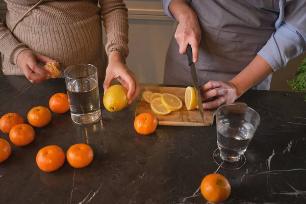 灰色のエプロンの認識できない女性は 妊娠中の友人が自宅で大理石のキッチンカウンターに立ってチョッピングボード上のレモンを切っている間に健康的なフルーツスナックを調理するのを助けます — ストック写真