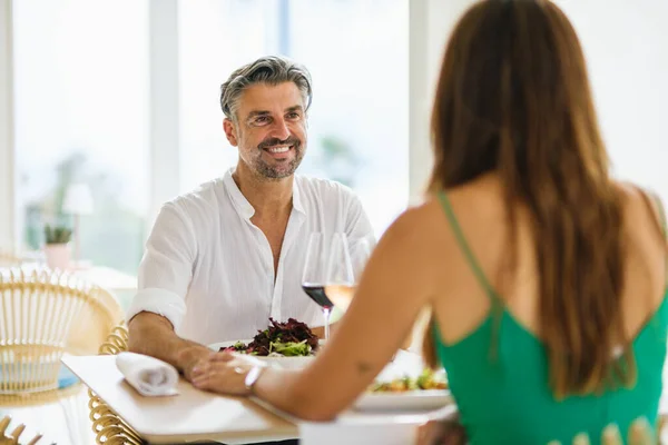 Loving couple in elegant clothes looking at each other with smile and holding hands while sitting at table with tasty salads and wine during date in restaurant on street