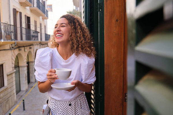 Positive young female with eyes closed laughing while drinking tea and standing on balcony over narrow city street