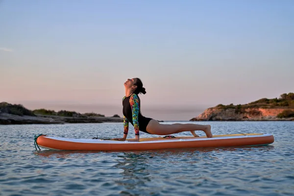 夜には青い海の中のパドルボード上でヨガを練習しながらウルドヴァ シャナサナポーズをとりながら水着姿の若い女性の側面図 — ストック写真