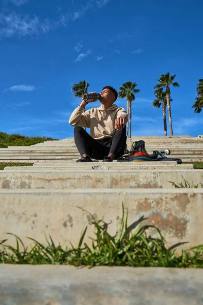 低角度ヒスパニック男のフルボディは 石の階段に座って 日当たりの良い都市公園でスケートボードで訓練した後 リフレッシュのために水を飲む — ストック写真