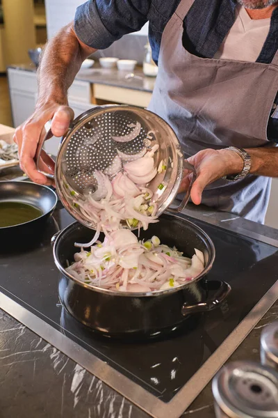 在现代厨房做饭的同时 匿名男性厨师将从菜刀上切下的洋葱倒进炉子上的油锅里 — 图库照片