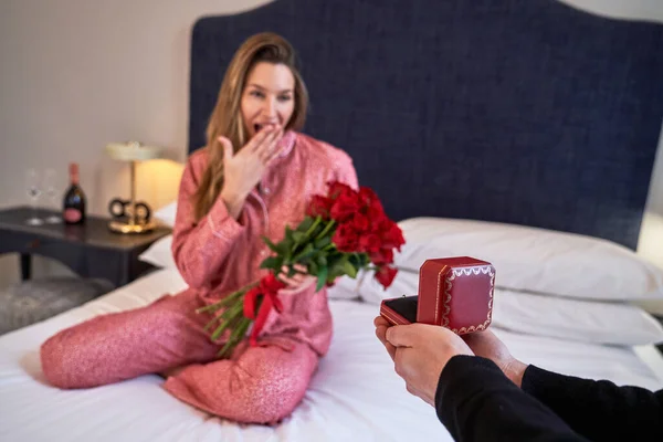 Arms of crop male giving gift box to surprised happy girlfriend with red roses in pajama on bed