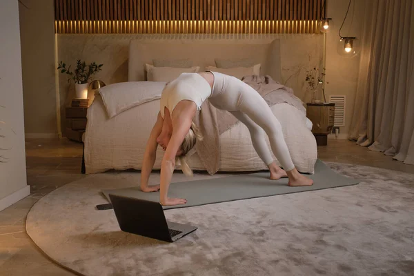 在Urdhva Dhanurasana 穿着运动服的全身上下都是柔韧的赤脚女子 她们在网上通过笔记本电脑进行瑜伽练习和伸展身体 同时保持平衡 — 图库照片