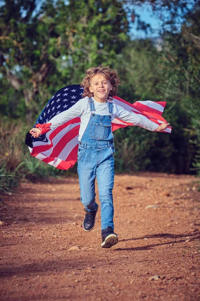 Bütün vücudu, yaz günü ulusal bir Amerikan bayrağıyla yolda koşarken kameraya bakan neşeli bir çocuk.