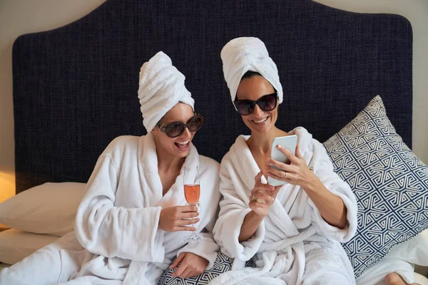 戴着太阳镜和毛巾头巾的快乐的女性朋友们穿着毛茸茸的浴袍 坐着一杯玫瑰香槟酒 一边用智能手机自拍 一边在宾馆的房间里度过周末 — 图库照片