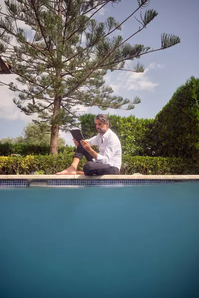 Dojrzały Mężczyzna Siedzi Brzegu Basenu Używa Tabletu Słoneczny Dzień Podwórku — Zdjęcie stockowe