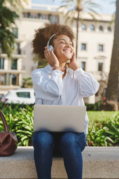 Uśmiechnięta Afroamerykanka Słuchawkach Pracująca Zdalnie Laptopie Słuchająca Muzyki Siedząc Kamiennej — Zdjęcie stockowe