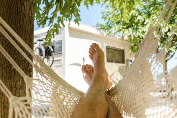 Crop barefoot man relaxing in hammock near tree trunk and caravan on sunny weekend day in summer