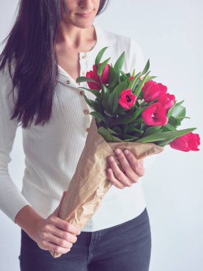 Beyaz arka planda kağıt ambalajda kırmızı lalelerle dikilen, uzun siyah saçlı, tanınmayan bir kadın.