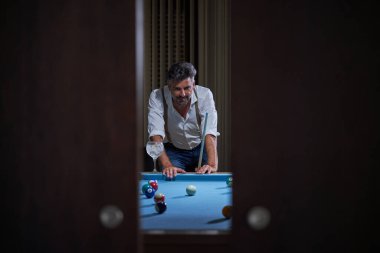 Mavi bilardo masasına yaslanmış, elinde bir bardak kokteyl olan ve karanlık oturma odasında bilardo oynarken bilardo toplarına bakan yetişkin bir adamın fotoğrafına bakın.