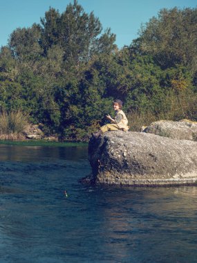 Yazın güneşli bir hafta sonu, çalı çırpı ve ağaçlara karşı sert kayalarda oturan ve balık tutan bir çocuk.