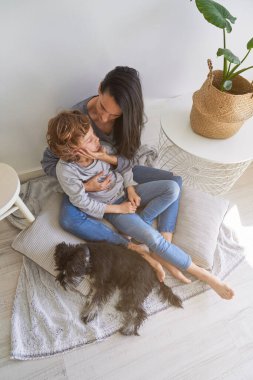 Huzurlu çocuk kıvırcık saçlı ve gözleri kapalı. Evde aydınlık bir odada otururken üstsüz annesini nazikçe kucaklıyor. 