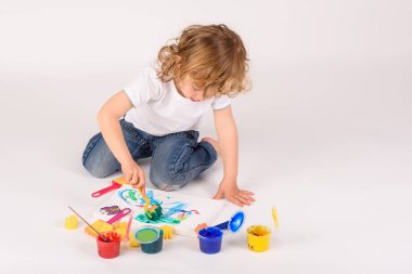 Işık stüdyosundaki beyaz arka planda renkli boyaların yanında kağıda boyanmış odaklanmış çıplak ayaklı bir çocuk.