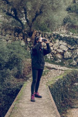Doğa yürüyüşü sırasında dar bir yolda duran dürbünle doğaya hayranlık duyarak gülümseyen koyu renk saçlı ve spor ayakkabılı genç aktif kadın gezginin tüm vücudu.