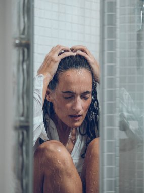 Beyaz gömlekli, koyu renk saçlı, üzgün bir kadın. Duşun altında oturmuş, saçlarına dokunuyor.