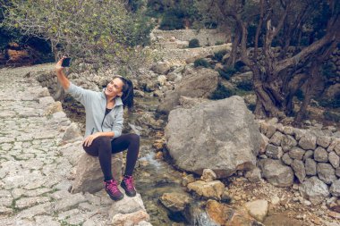 Tüm vücudu, Mallorca Soller Biniaraix 'de akarsu deresinin kenarında otururken cep telefonuyla fotoğraf çeken mutlu bir kadın.