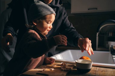 Sıradan giysiler içinde komik küçük bir çocuk ve taze olgun portakalları soyan şapkalı bir çocuğun mutfakta ekin yetiştiren isimsiz bir babayla yan görünüşü.