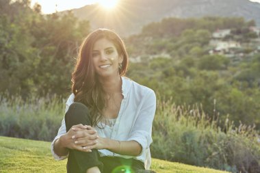 Neşeli genç bayan, güneşli bir yaz günü bahçenin yakınındaki çimenlikte dinlenirken gülümseyerek kameraya bakıyor.