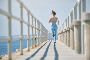 Anonim fit kadın koşucunun, spor kıyafetleri içinde güneş ışığında deniz üzerinde çitli köprüde koşuşunun arka görüntüsü.