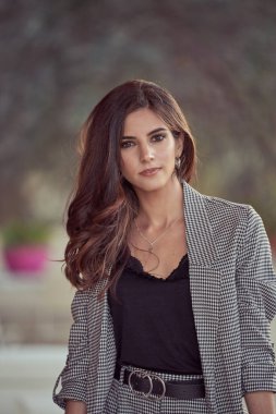 Zarif kıyafetli, siyah kıvırcık saçlı çekici bir kadın sokakta bulanık arka planda kameraya bakıyor.