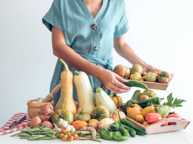 Tanımlanamayan bir kadın elbise giymiş, masanın üzerine sarımsak ve bezelye koyarak ve domatesleri beyaz duvara dayayarak taze balkabağı, biber ve soğan yığının yanında duruyor.