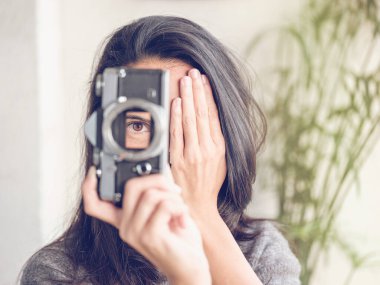Tanımlanamayan uzun siyah saçlı bir kadın eski film kamerasından bakarken yüzünün yarısını eliyle kapatıyor ve fotoğraf çekiyor.