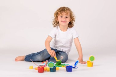 Stüdyoda beyaz arka planda guaj boyaları çizerken kameraya bakan neşeli bir çocuk vücudu.