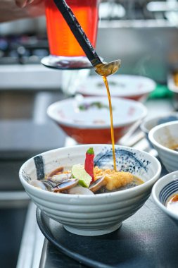 Anonim bir aşçı seramik kaseye yağlı sos ekliyor. Erişte çorbası, çıtır tavuk derisi midye, kırmızı biber ve limon dilimleri ile restoranda sipariş vermek üzere servis ediliyor.