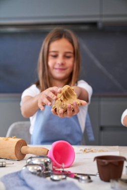 Önlüklü iyimser kız apartmanın açık mutfağında çeşitli malzemelerle dağınık masada dururken kurabiye hamuru yoğuruyor.