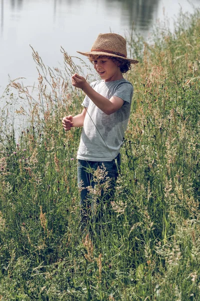 夏の日の穏やかなチャンネルに立って 離れて見ている間 背の高い草で遊ぶわら帽子とジーンズの陽気な子供 — ストック写真