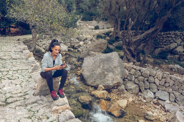 在Mallorca Soller Biniaraix 全身上下都是笑容满面的女旅行者 她们坐在瀑布溪边的石头路上 还在浏览手机 — 图库照片