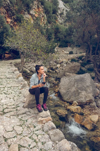 侧观全景全景心满意足的女人 一边坐在岩石上 一边拿着智能手机 一边望着远方 一边享受着山区溪流溪流边的旅行 — 图库照片