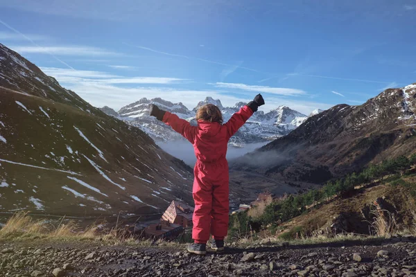 赤いアウターウェアで認識できない子供が腕を上げ 村と谷の近くに立って雪山を鑑賞しながら自由を楽しむバックビュー — ストック写真