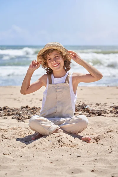 Full body of happy child in jumpsuit touching straw hat and looking at camera while enjoying summer holidays at seaside