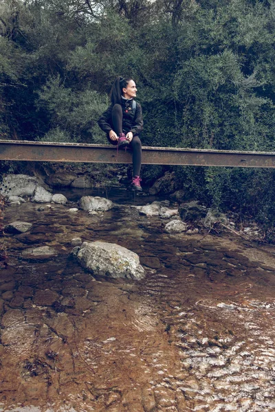 穿着休闲装的女游客坐在马洛卡森林河畔的石桥上 望着远方 — 图库照片