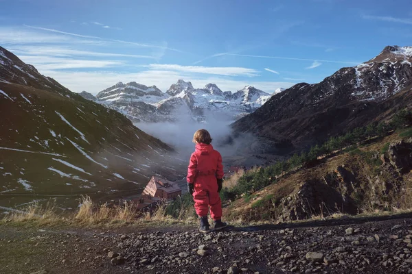 丘の斜面に立っている赤いアウターウェアの子供のバックビューと春に雪山で時間を過ごしながら谷の家を賞賛 — ストック写真