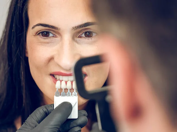 Dark haired lady looking at camera while crop blurred dentist selecting hue of teeth with loupe and shade scale