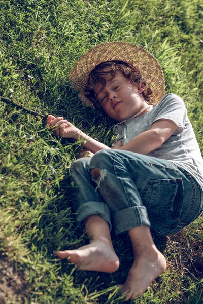 フルボディの上から かわいい裸足の小僧 ウィッカー帽子とジーンズ 緑の草の上に横たわり 日光の中で眠る — ストック写真