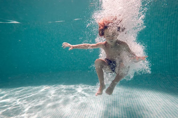 青い壁にジャンプしながらプールの清潔な透明な水の下に突っ込む目を閉じた裸足の子供の全身 — ストック写真