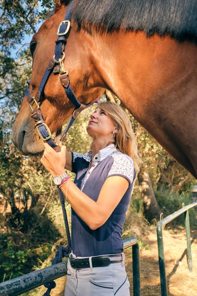 Content female equestrian caressing muzzle of chestnut obedient horse in harness while standing on ranch in summer